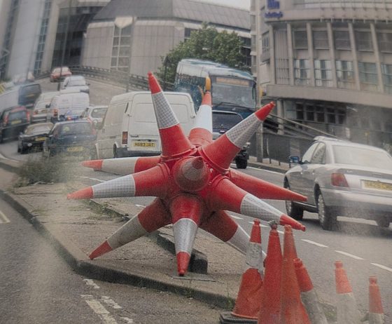 Banksy's traffic-cone sculpture - eight red-and-white traffic cones welded into a giant star and left on a central reservation in London traffic, photographed from a book page